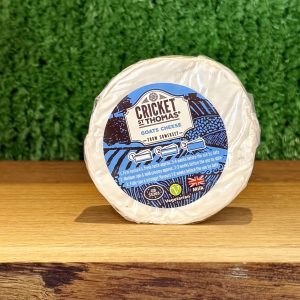 A picture of a small, round 100g Cricket St Thomas goats' cheese. Is is on a wooden board with a green background