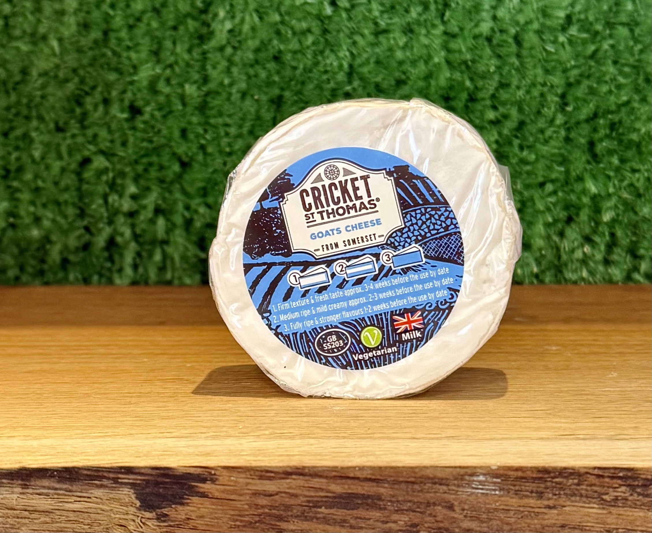 A picture of a small, round 100g Cricket St Thomas goats' cheese. Is is on a wooden board with a green background