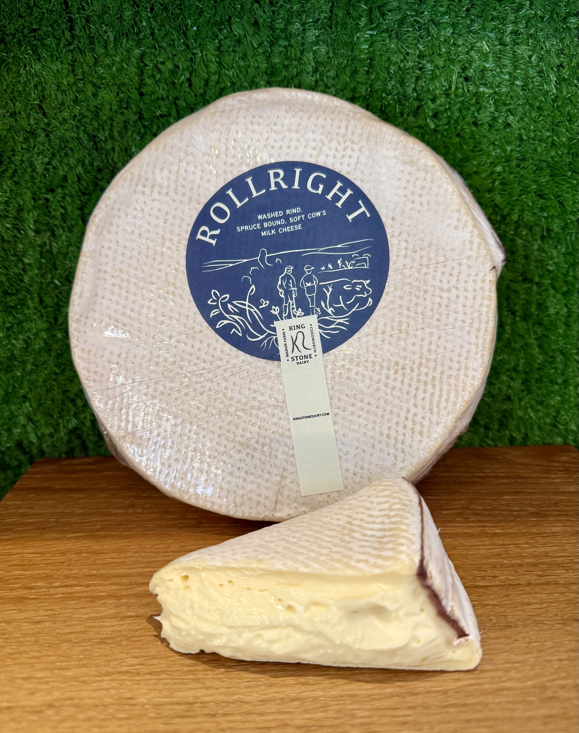 A picture showing a wheel of Rollright cheese with a cut wedge next to it. They are positioned on a wooden board with a green backdrop