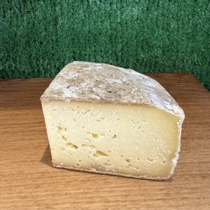 A picture of a wedge of Tomme des Alpes cheese. It is sitting on a wooden board with a green background