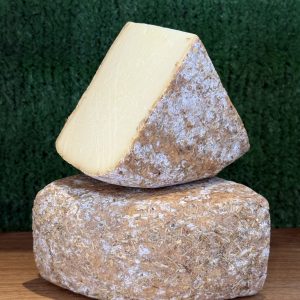 A photo showing 2 wedges of Witheridge cheese. One is balanced on the other at a jaunty angle. They are positioned on a wooden board with a green background