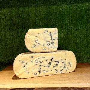 A picture of half a Kingcott Blue cheese with a quarter piece sat on top of it. It is on a wooden board with a green background