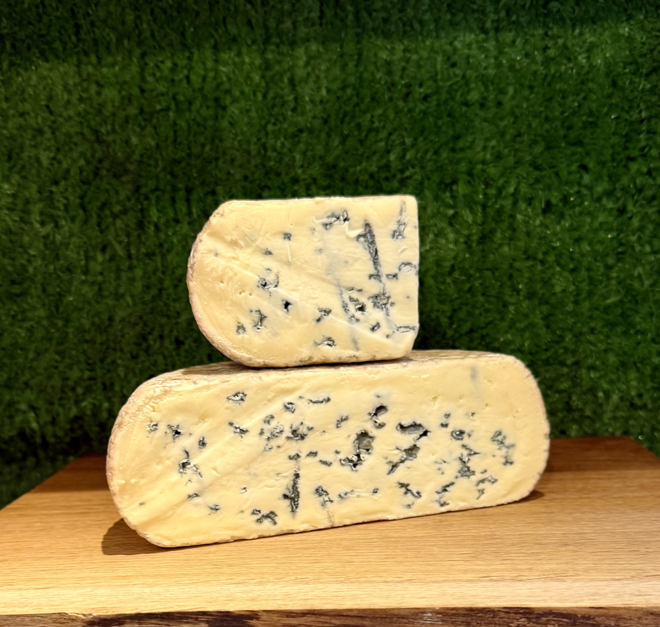 A picture of half a Kingcott Blue cheese with a quarter piece sat on top of it. It is on a wooden board with a green background
