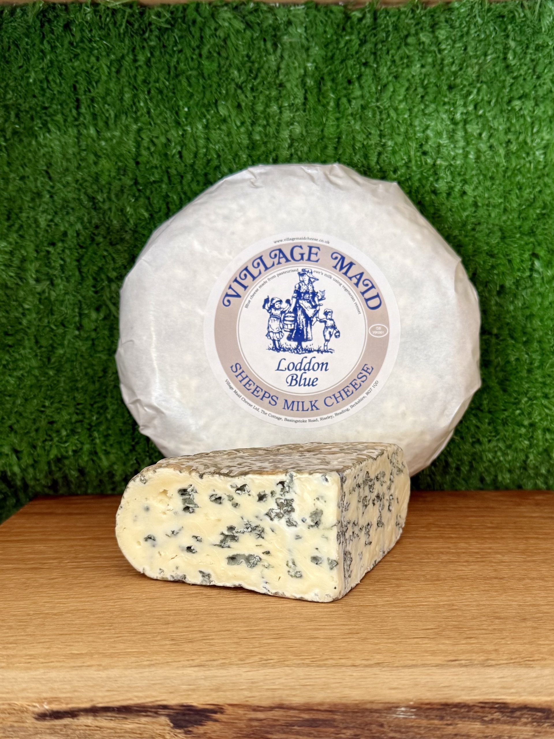 A picture showing a wedge of Loddon Blue. It is sat on a wooden board with a whole round of cheese propped up behind it