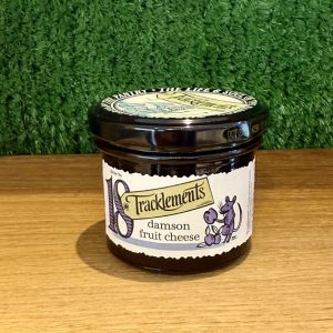 A picture of a jar of Tracklememts Damson Fruit Cheese. It is positioned on a wooden board with a green background.