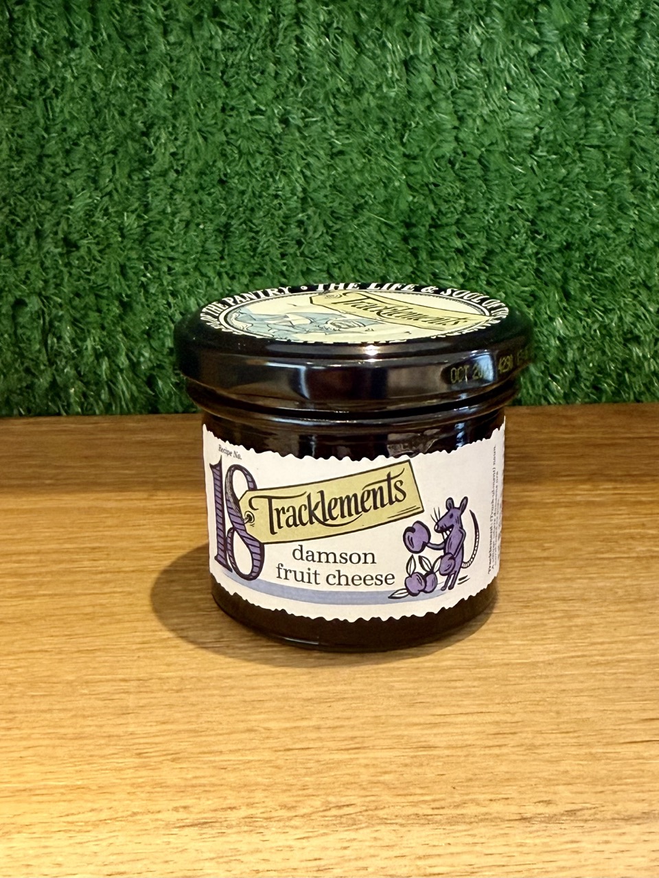 A picture of a jar of Tracklememts Damson Fruit Cheese. It is positioned on a wooden board with a green background.