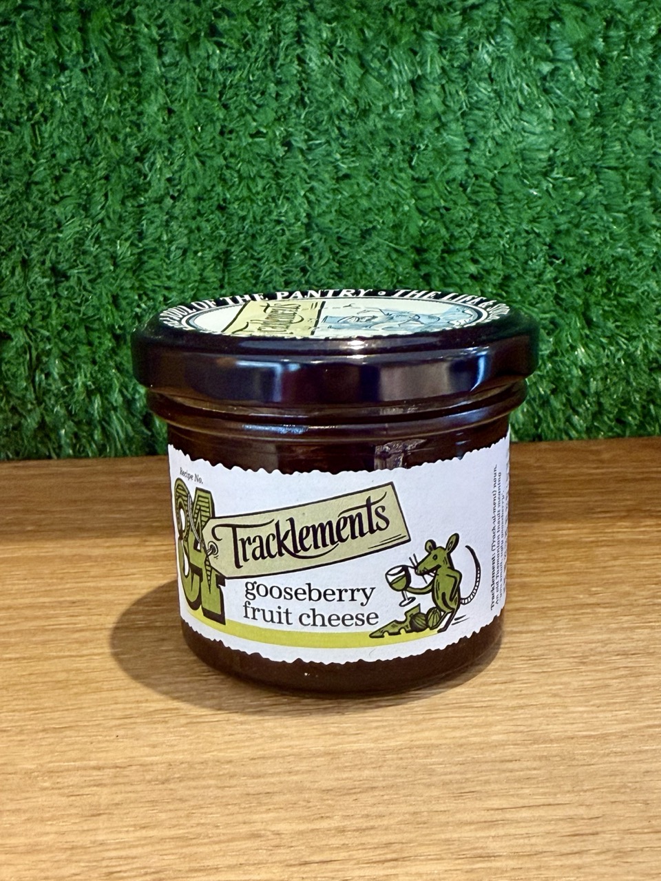 A picture of a jar of Tracklments Gooseberry Fruit Cheese. It is shown on a wooden board with a green background