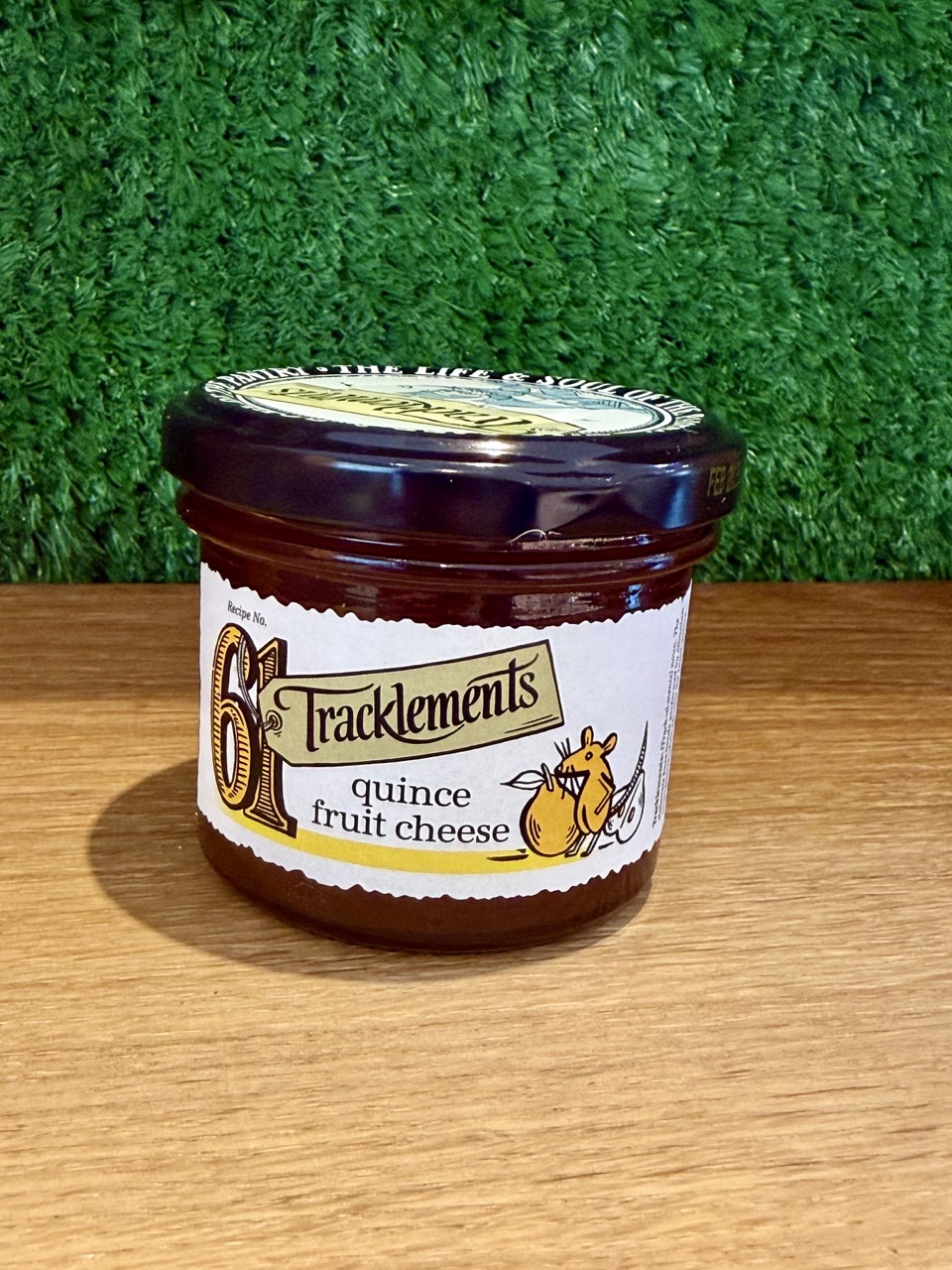 A picture of a jar of Tracklements Quince Fruit Cheese. It is positioned on a wooden board with a green background