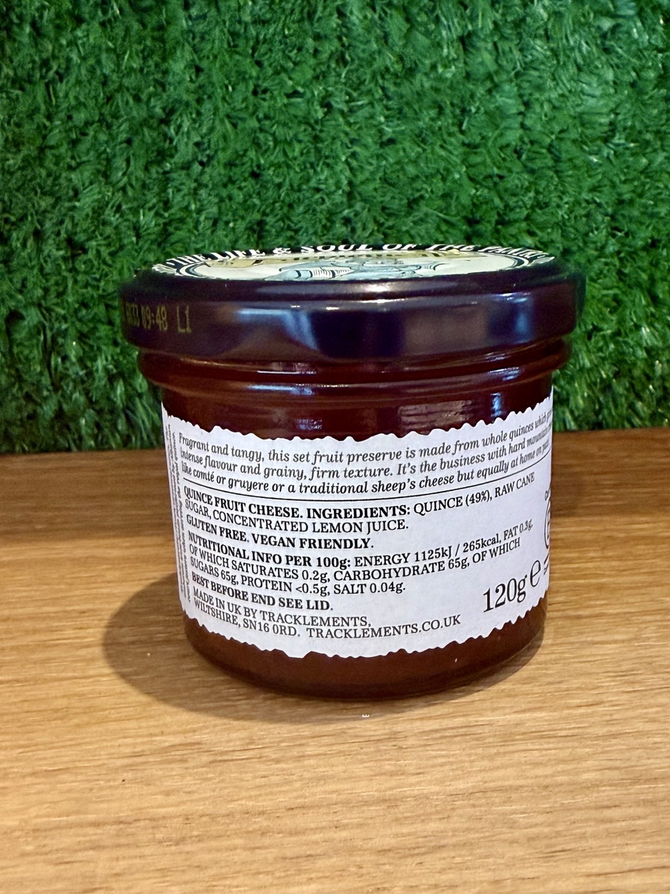 A picture of the back of a jar of Tracklements Quince Fruit Cheese. It is positioned on a wooden board with a green background