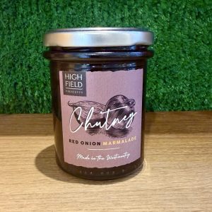 A picture of a jar of Red Onion Marmalade. It is positioned on a wooden board with a green background