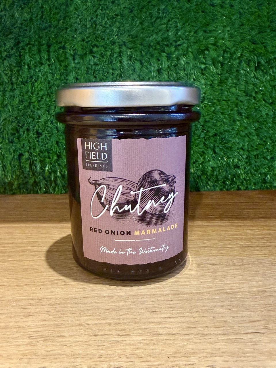 A picture of a jar of Red Onion Marmalade. It is positioned on a wooden board with a green background