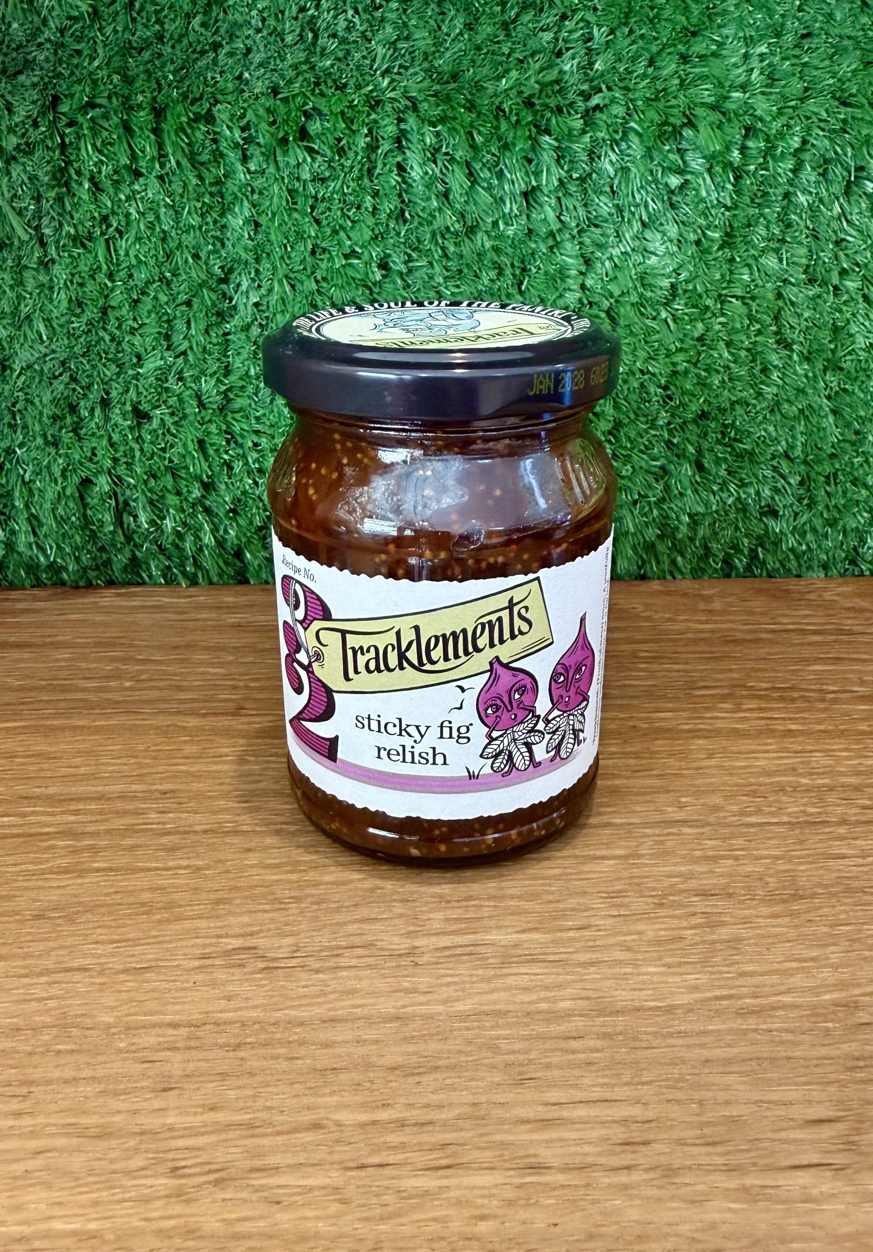 A jar of Sticky Fig Relish. It is pictured on a wooden board with a green background