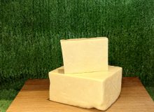 A picture showing 2 pieces of Barber's 1833 Vintage Reserve Cheddar. They are positioned on a wooden board with a green background