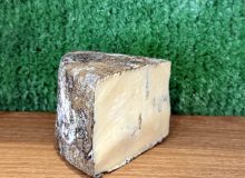 A picture of a wedge of Bidlea Blue cheese. It has been placed on a wooden board with a green backdrop