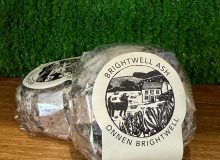 A picture showing two Brightwell Ash cheeses. They are positioned on a wooden board with a green background