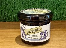 A picture of a jar of Tracklememts Damson Fruit Cheese. It is positioned on a wooden board with a green background.