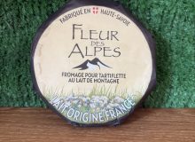 A picture of a wheel of Fleur des Alpes cheese. It is on a wooden board with a green background