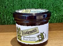 A picture of a jar of Tracklments Gooseberry Fruit Cheese. It is shown on a wooden board with a green background