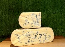 A picture of half a Kingcott Blue cheese with a quarter piece sat on top of it. It is on a wooden board with a green background