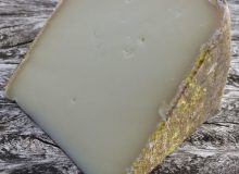 A photograph depicting a wedge of Ossau-Iraty PDO cheese. It has been positioned on a rustic wooden table.