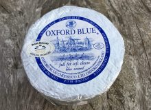 A picture of an Oxford Blue Mini Cheese. It is wrapped in foil and is positioned on a wooden table