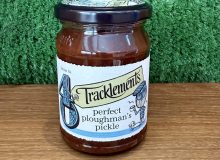 A photo of a jar of Perfect Ploughman's Pickle. It is pictured on a wooden board with a green background
