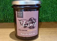 A picture of a jar of Red Onion Marmalade. It is positioned on a wooden board with a green background