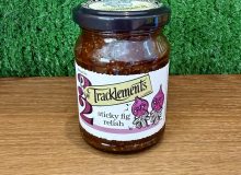 A jar of Sticky Fig Relish. It is pictured on a wooden board with a green background