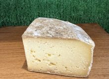 A picture of a wedge of Tomme des Alpes cheese. It is sitting on a wooden board with a green background