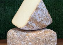 A photo showing 2 wedges of Witheridge cheese. One is balanced on the other at a jaunty angle. They are positioned on a wooden board with a green background