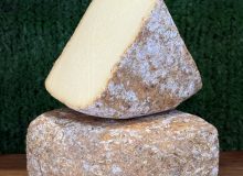 A photo showing 2 wedges of Witheridge cheese. One is balanced on the other at a jaunty angle. They are positioned on a wooden board with a green background