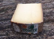 A picture of a slice of Aged Comté PDO cheese. It is positioned on a rustic looking wooden table