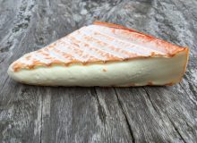 A picture of a wedge of Brebirousse d'Argental Cheese. It has been placed on a rustic, wooden table and is looking ripe and ready to eat!