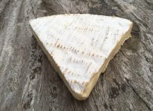 A wedge of Dongé Brie is depicted on a rustic looking wooden table.