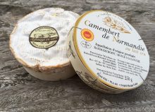 A picture of a whole Camembert de Normandie PDO. It is shown with its lid resting on its rim, whilst sitting on a rustic wooden table.