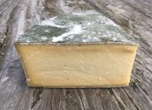 A picture showing a cut wedge of Cornish Yarg Cheese. It is sat on a rustic wooden table and it's nettle covered rind looks great