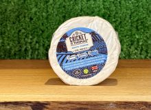 A picture of a small, round 100g Cricket St Thomas goats' cheese. Is is on a wooden board with a green background