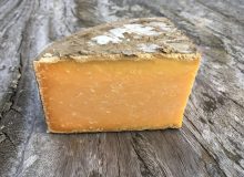 A photo depicting a wedge of Double Gloucester Cheese. It is positioned on a rustic, wooden table