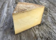 A photo of a wedge of Duckett's Caerphilly Cheese. It is positioned on a rustic wooden table.