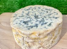 A picture of half a cylinder of Fourme d'Ambert PDO Cheese. It is on a wooden board with a green backdrop