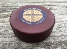 A picture of a round of Godminster Cheddar Cheese. It is positioned on a rustic, wooden table