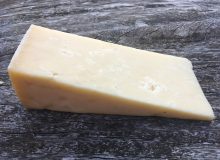 A picture of a wedge of Isle of Mull Cheddar Cheese. It is positioned on a rustic, wooden table.
