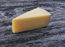 A photograph of a wedge of Lincolnshire Poacher cheese. It is sitting on a rustic, wooden table and looks ready to eat!