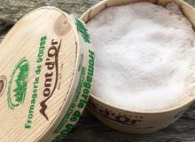 A picture of a Vacherin Mont d'Or Baby cheese. It is in its box with the lid off and has been placed on a rustic wooden table