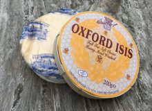 A photo of a whole Oxford Isis Baby cheese. The lid of its box is half off, exhibiting the cheese within. It has been placed on a rustic looking wooden table.