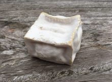 A picture of a Pave D'Affinois Cheese. It is a white cube which is starting to sag. It has been placed on a rustic wooden table.