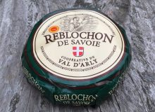 A photo of a whole Reblochon PDO cheese. It has been placed on a rustic wooden table.