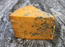 A picture of a wedge of Shropshire Blue cheese sitting on a wooden table. It is orange in colour with blue veining throughout