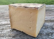 A photograph of a wedge of Single Gloucester Cheese. It is pictured on a rustic, wooden table