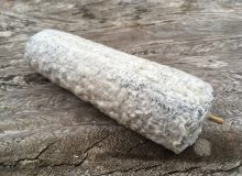 A photograph of a whole Sainte-Maure de Touraine PDO cheese. It is shown on a rustic wooden table